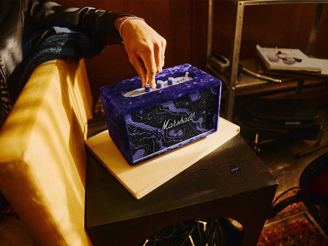 Détail rapproché de l'enceinte Marshall Acton III Hendrix Edition montrant le revêtement en velours violet écrasé à motif cosmic swirl, le panneau de contrôle supérieur en argent brossé avec ses trois boutons rotatifs violets et son logo Marshall en script argenté sur la grille frontale, une main aux bagues argentées s'apprêtant à actionner le bouton central, dans une mise en scène studio aux tonalités chaudes