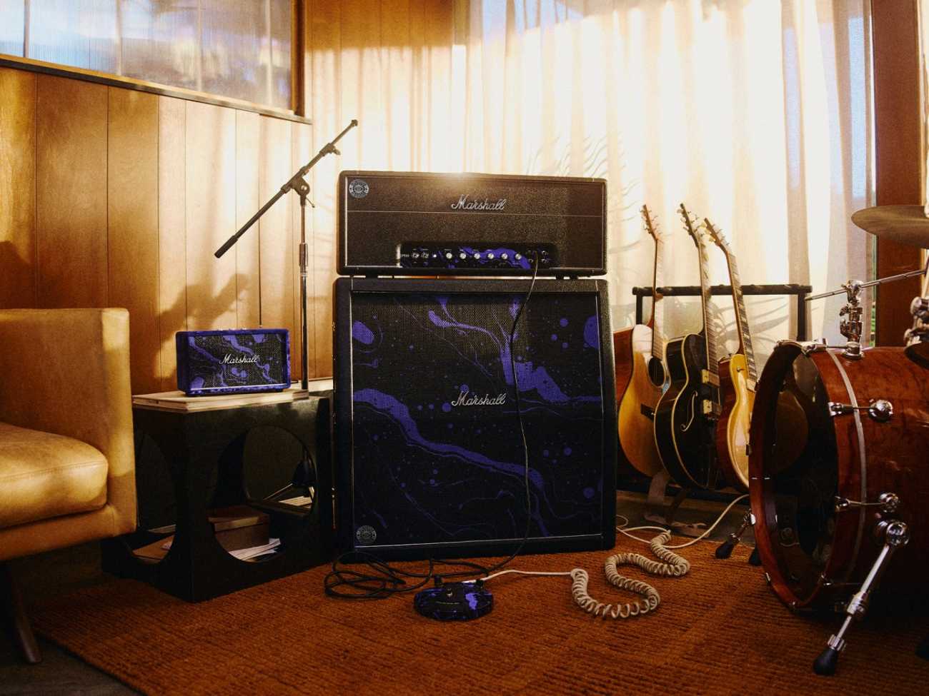 Mise en scène studio de la collection complète Marshall x Hendrix 60th Anniversary, avec la tête d'ampli 1959 Handwired posée sur le baffle 1960 AJMH 4x12 en finition cosmic swirl noir et violet, l'enceinte Bluetooth Acton III Hendrix Edition en velours violet sur table basse à gauche, et la pédale Fuzz Face Dunlop édition limitée au sol, dans une pièce avec guitares acoustiques et batterie en arrière-plan