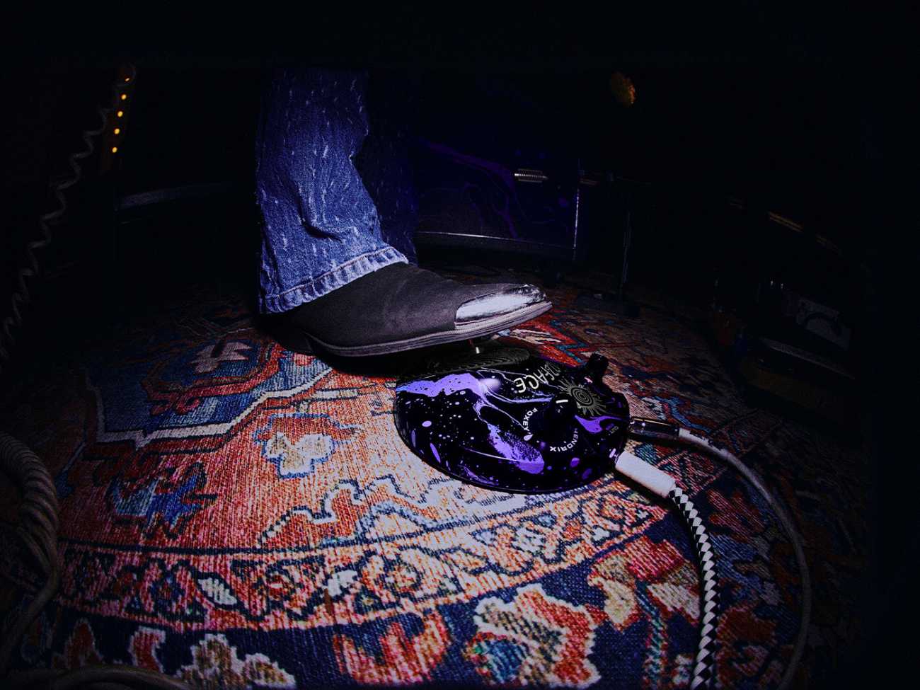 Pédale de distorsion Fuzz Face Dunlop édition limitée Marshall x Hendrix 60th Anniversary, en finition cosmic swirl noir et violet assortie à la collection, photographiée au sol sur un tapis persan rouge et bleu avec un pied chaussé d'une santiag noire et bleu jean s'apprêtant à l'enclencher, câble jack tressé noir et blanc connecté à la sortie, ambiance scénique faiblement éclairée