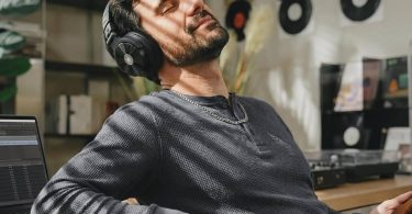 Homme barbu en pull gris portant le casque OneOdio Studio Max 2 noir aux coques aluminium brossé, écoutant de la musique en Bluetooth LDAC depuis son smartphone dans un home studio avec MacBook ouvert sur une DAW et étagère de vinyles en arrière-plan