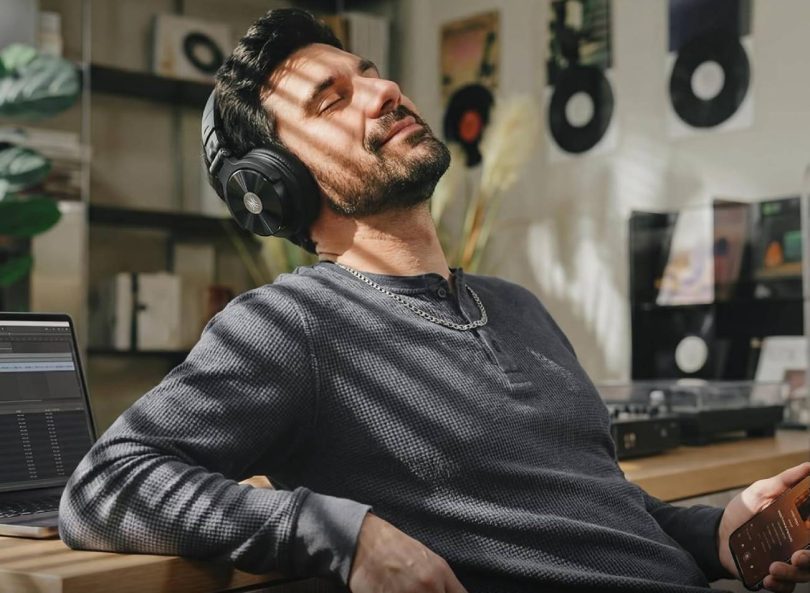 Homme barbu en pull gris portant le casque OneOdio Studio Max 2 noir aux coques aluminium brossé, écoutant de la musique en Bluetooth LDAC depuis son smartphone dans un home studio avec MacBook ouvert sur une DAW et étagère de vinyles en arrière-plan
