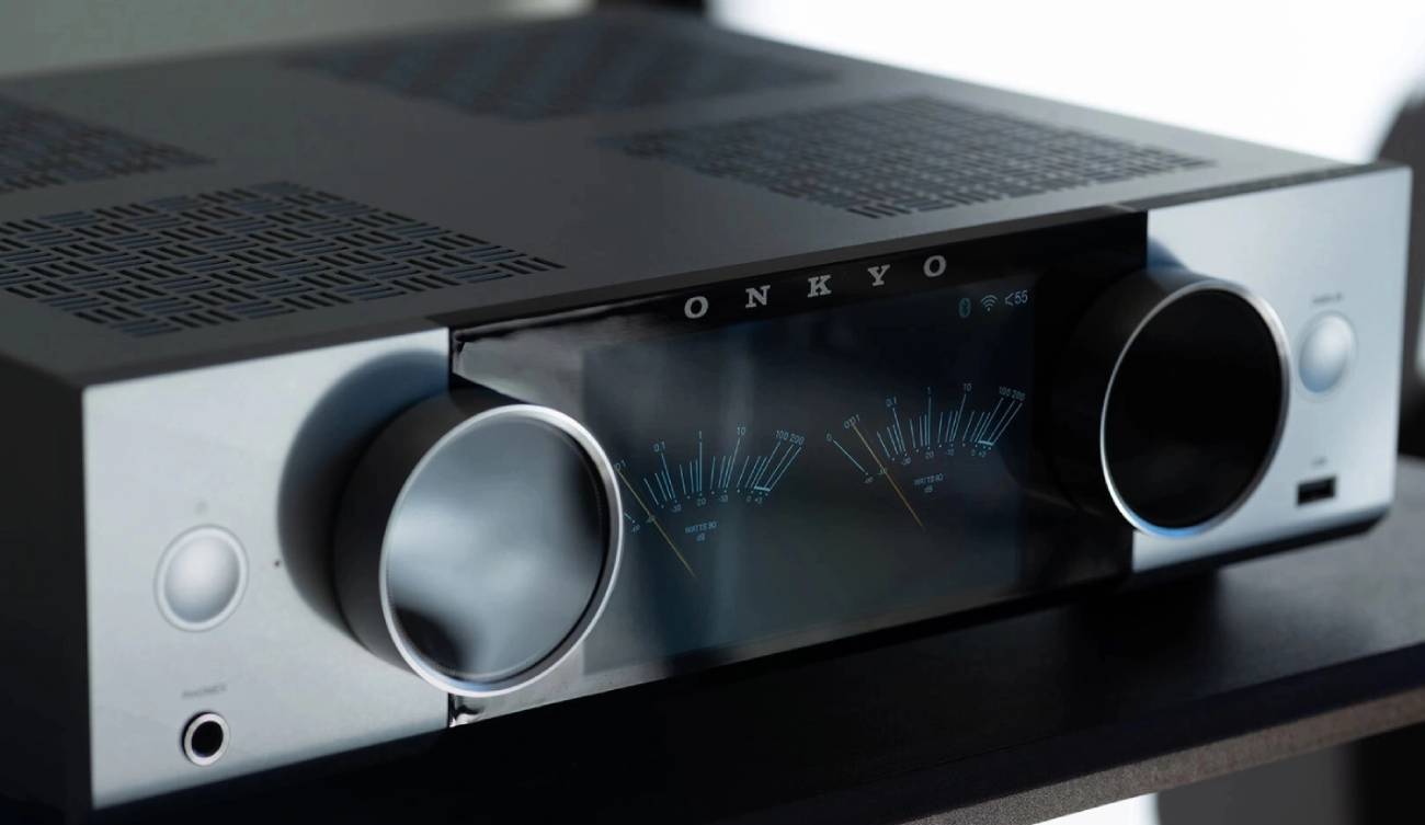 Façade de l'amplificateur Onkyo Muse, écran couleur affichant deux vumètres analogiques virtuels gradués en watts avec icônes Bluetooth et Wi-Fi, logo ONKYO, deux grosses molettes rotatives, port USB-A frontal et prise casque jack 6,35 mm, capot ajouré au motif San Kuzushi