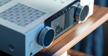 Gros plan sur l'amplificateur Onkyo Muse en finition argent : écran couleur LCD 5,46 pouces affichant une pochette d'album, logo ONKYO, deux molettes rotatives en aluminium, prise casque jack 6,35 mm en façade et grille de ventilation supérieure au motif japonais San Kuzushi