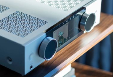 Gros plan sur l'amplificateur Onkyo Muse en finition argent : écran couleur LCD 5,46 pouces affichant une pochette d'album, logo ONKYO, deux molettes rotatives en aluminium, prise casque jack 6,35 mm en façade et grille de ventilation supérieure au motif japonais San Kuzushi