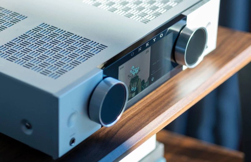 Gros plan sur l'amplificateur Onkyo Muse en finition argent : écran couleur LCD 5,46 pouces affichant une pochette d'album, logo ONKYO, deux molettes rotatives en aluminium, prise casque jack 6,35 mm en façade et grille de ventilation supérieure au motif japonais San Kuzushi