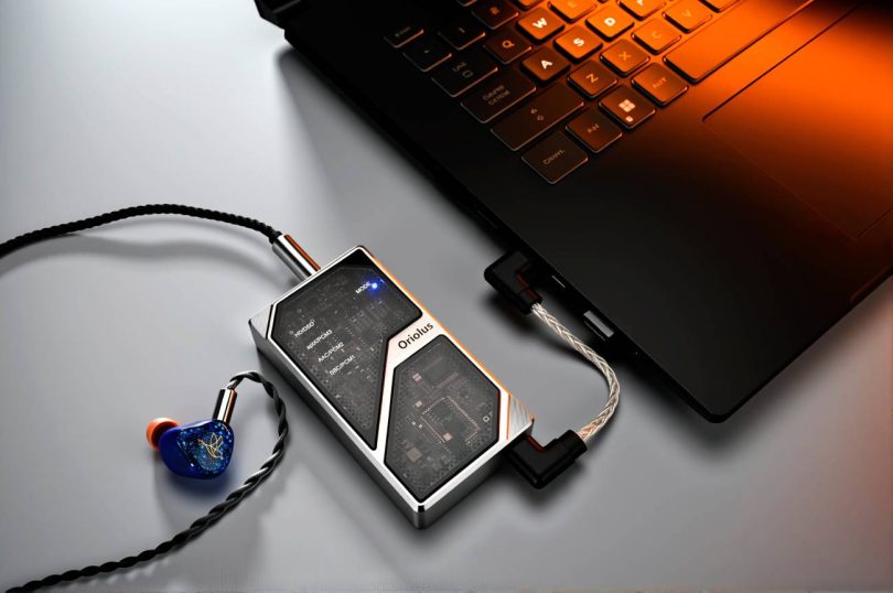 DAC portable Oriolus 1795MKII AL en aluminium avec panneau de verre semi-transparent laissant apparaître le circuit imprimé interne, connecté à un laptop via câble USB-C coudé et à un IEM bleu via sortie 4.4mm balanced à câble tressé argenté