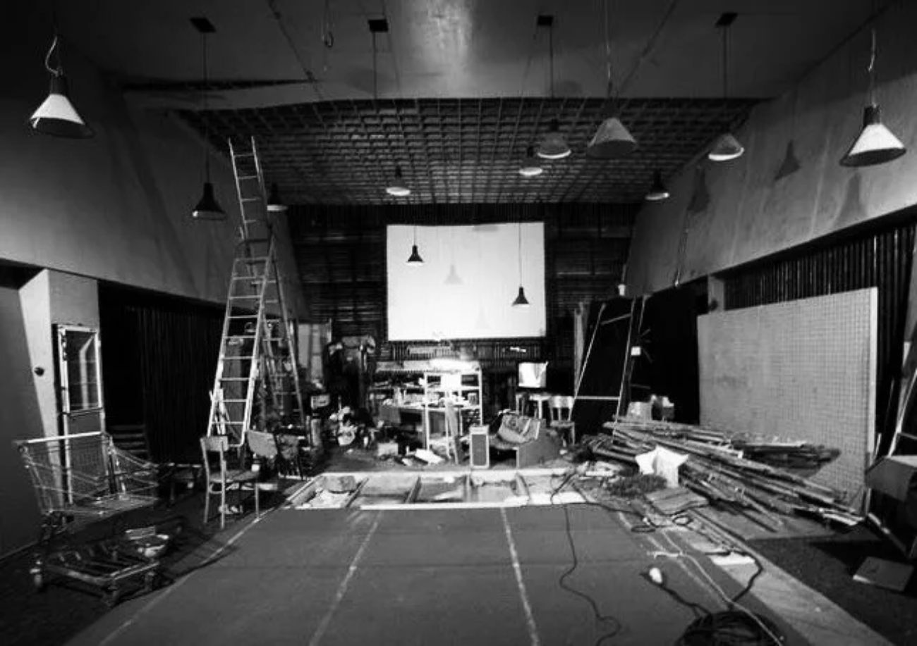 Vue intérieure d'un plateau Foley traditionnel en noir et blanc : grand studio fermé avec écran de projection central pour synchronisation image, suspensions de spots, échelles, matériaux et accessoires divers éparpillés au sol pour la production de bruitages indoor