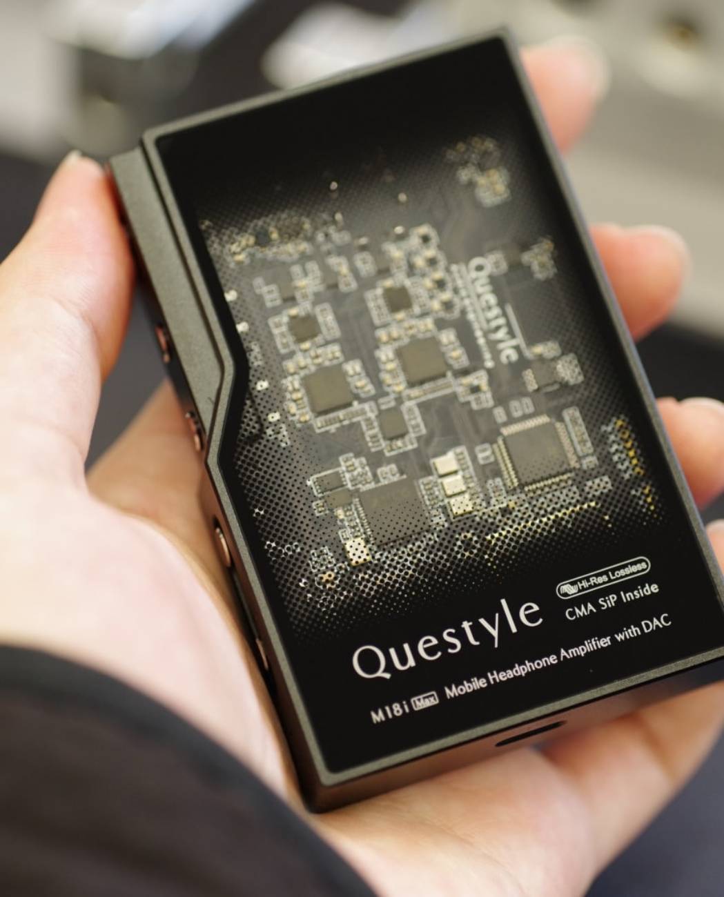 Vue rapprochée du Questyle M18i MAX tenu en main, dos en verre semi-transparent qui dévoile le PCB et les modules SiP Current Mode Amplification brevetés Questyle, avec inscriptions Hi-Res Lossless, CMA SiP Inside, M18i Max Mobile Headphone Amplifier with DAC, sorties casque 3,5 mm et 4,4 mm visibles sur le côté gauche du boîtier