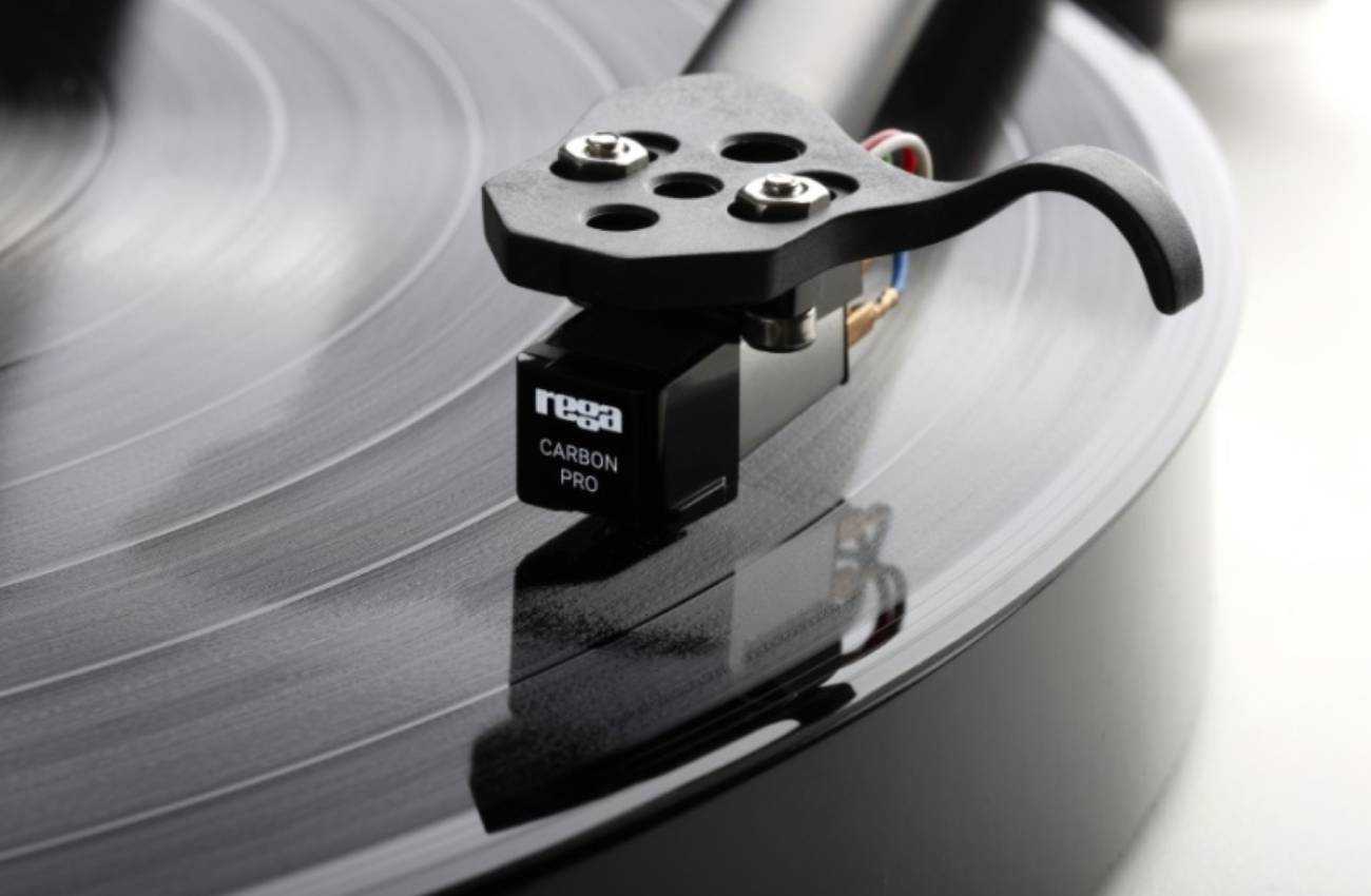 Vue de dessus en gros plan de la cellule Rega Carbon Pro avec marquage logo rega et inscription CARBON PRO sur la face avant, montée sur le headshell trois trous d'un bras de lecture Rega RB220, en cours de lecture sur un disque vinyle noir en rotation avec reflet de la cellule sur la surface du sillon