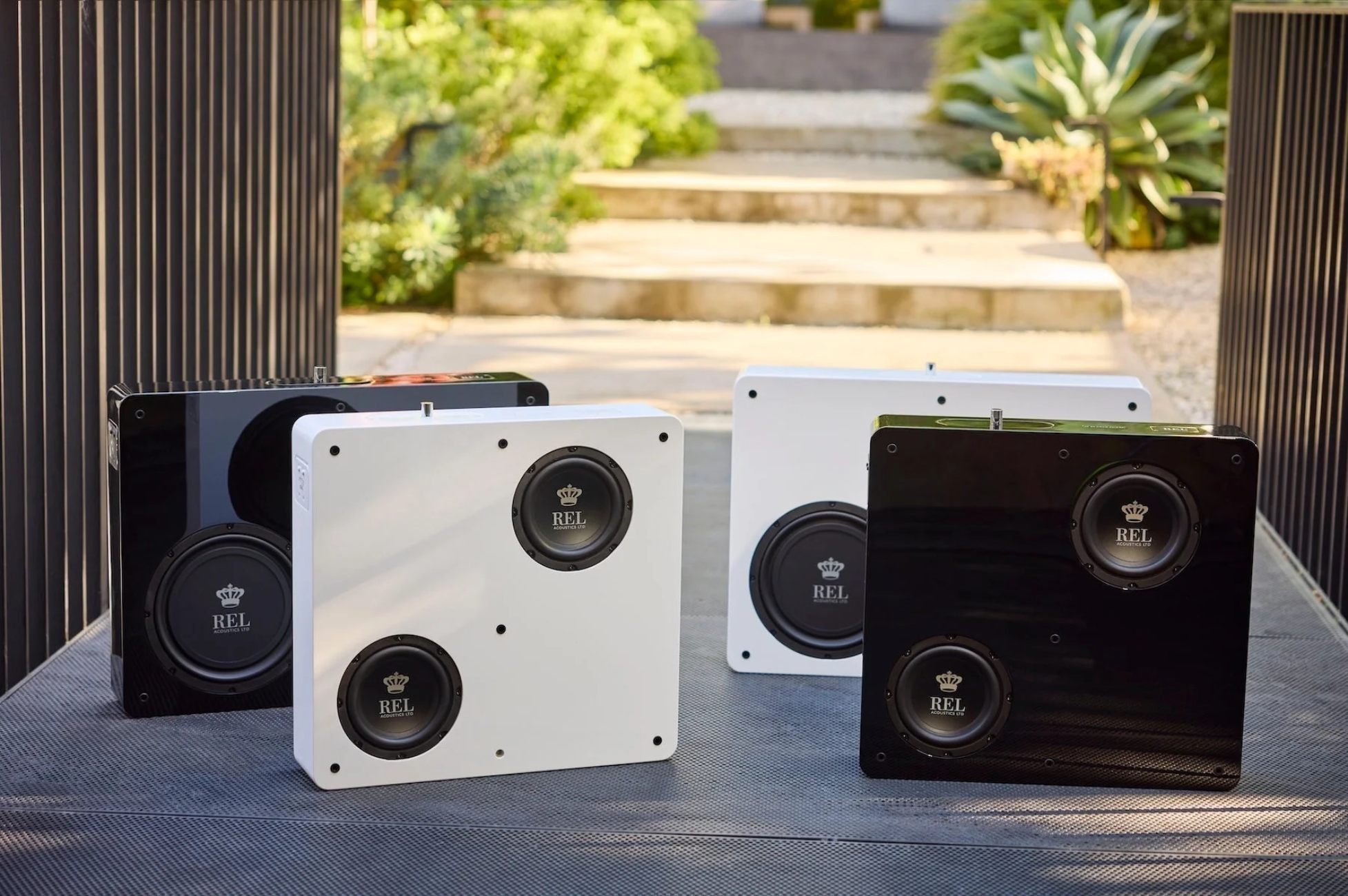 Famille complète de la série REL Planar avec les quatre modèles présentés sans grille acoustique : PL-1 et PL-2 en finitions noir laqué et blanc, drivers FibreAlloy et FlatPiston visibles, mise en scène extérieure jardin contemporain