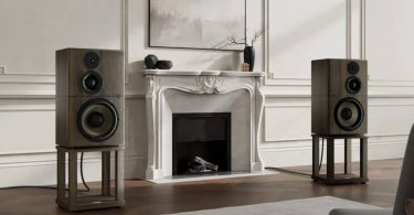 Paire d'enceintes Revival Audio Atalante Grande Réserve en finition chêne fumé, posées sur leurs stands dédiés en bois, encadrant une cheminée en marbre blanc dans un salon classique français avec moulures murales, parquet en chevrons et tableau abstrait au-dessus de la cheminée