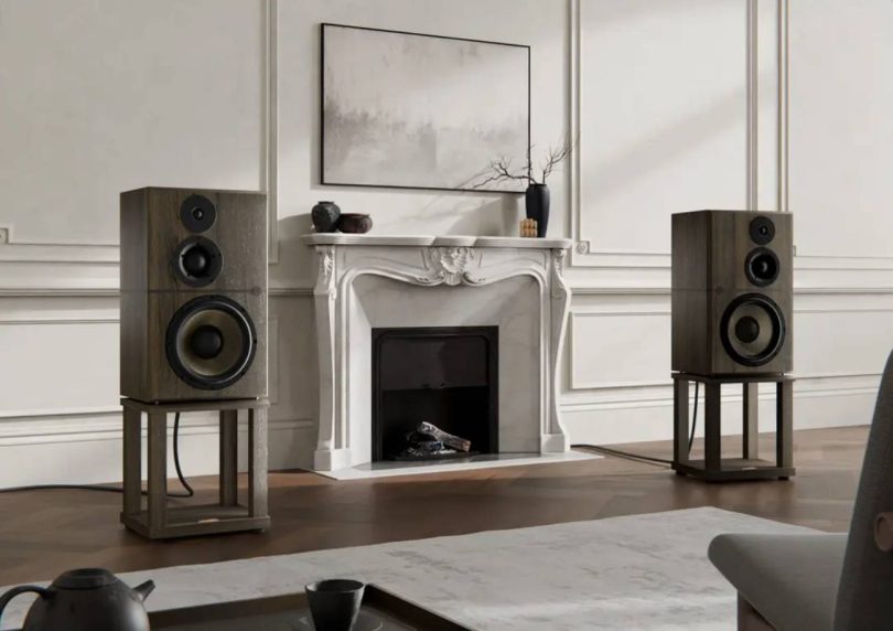 Paire d'enceintes Revival Audio Atalante Grande Réserve en finition chêne fumé, posées sur leurs stands dédiés en bois, encadrant une cheminée en marbre blanc dans un salon classique français avec moulures murales, parquet en chevrons et tableau abstrait au-dessus de la cheminée