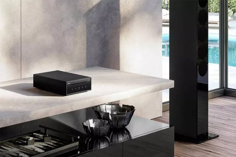 Lecteur CD Revox Studiomaster CD100 en finition noire posé sur une console en marbre clair dans un intérieur design contemporain, avec colonne Revox visible à droite et piscine en arrière-plan