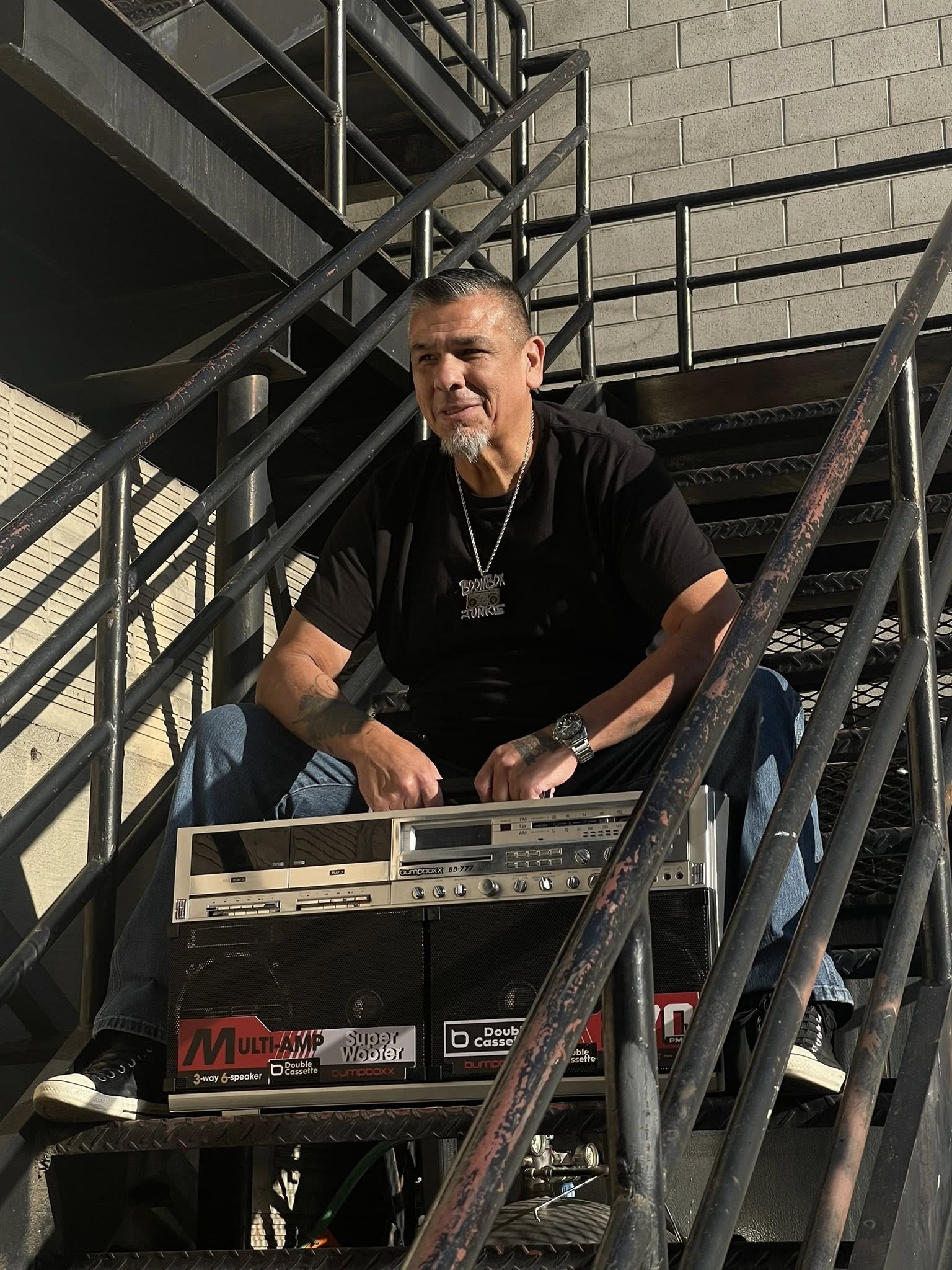 Rob Owens, fondateur de Bumpboxx, assis sur des escaliers métalliques extérieurs avec le boombox BB-777 posé devant lui. Le ghetto-blaster argenté affiche ses imposants haut-parleurs et sa double platine cassette sous la lumière du soleil.