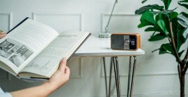Sangean DDR-8 radio portable DAB+ FM-RDS Bluetooth 5.2 en finition bois clair et grille champagne, posée sur table blanche avec vase céramique et fleurs séchées, écran OLED affichant heure 09:45 en grands chiffres style flip-clock, bouton rotatif central métal brossé