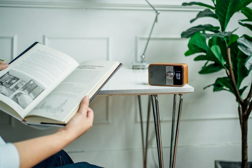Sangean DDR-8 radio portable DAB+ FM-RDS Bluetooth 5.2 en finition bois clair et grille champagne, posée sur table blanche avec vase céramique et fleurs séchées, écran OLED affichant heure 09:45 en grands chiffres style flip-clock, bouton rotatif central métal brossé
