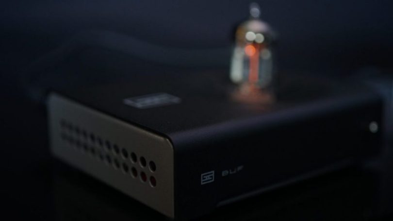 Tube buffer Schiit Buf en chassis noir mat avec côté champagne perforé, logo Schiit et marquage BUF, tube 6N1P rougeoyant visible à l'arrière en bokeh, ambiance studio audiophile sombre