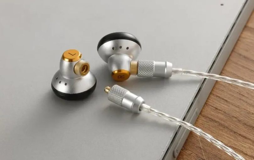 Paire d'écouteurs Sivga M260 en finition aluminium argenté avec accents en laiton doré, posés sur un bureau gris clair en bordure d'un plan de travail bois clair. La photo détaille la coque flathead façon vintage, le grillage frontal exposé, le connecteur MMCX détachable de l'enceinte gauche et le câble argenté tressé 4N OFC plaqué argent.