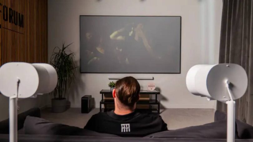 Configuration home cinéma Sonos en situation : barre de son Arc Ultra et caisson Sub 4 sous un grand téléviseur, encadrés par deux enceintes Era 300 blanches montées sur pieds en position surround, dans un salon contemporain pour une écoute Dolby Atmos