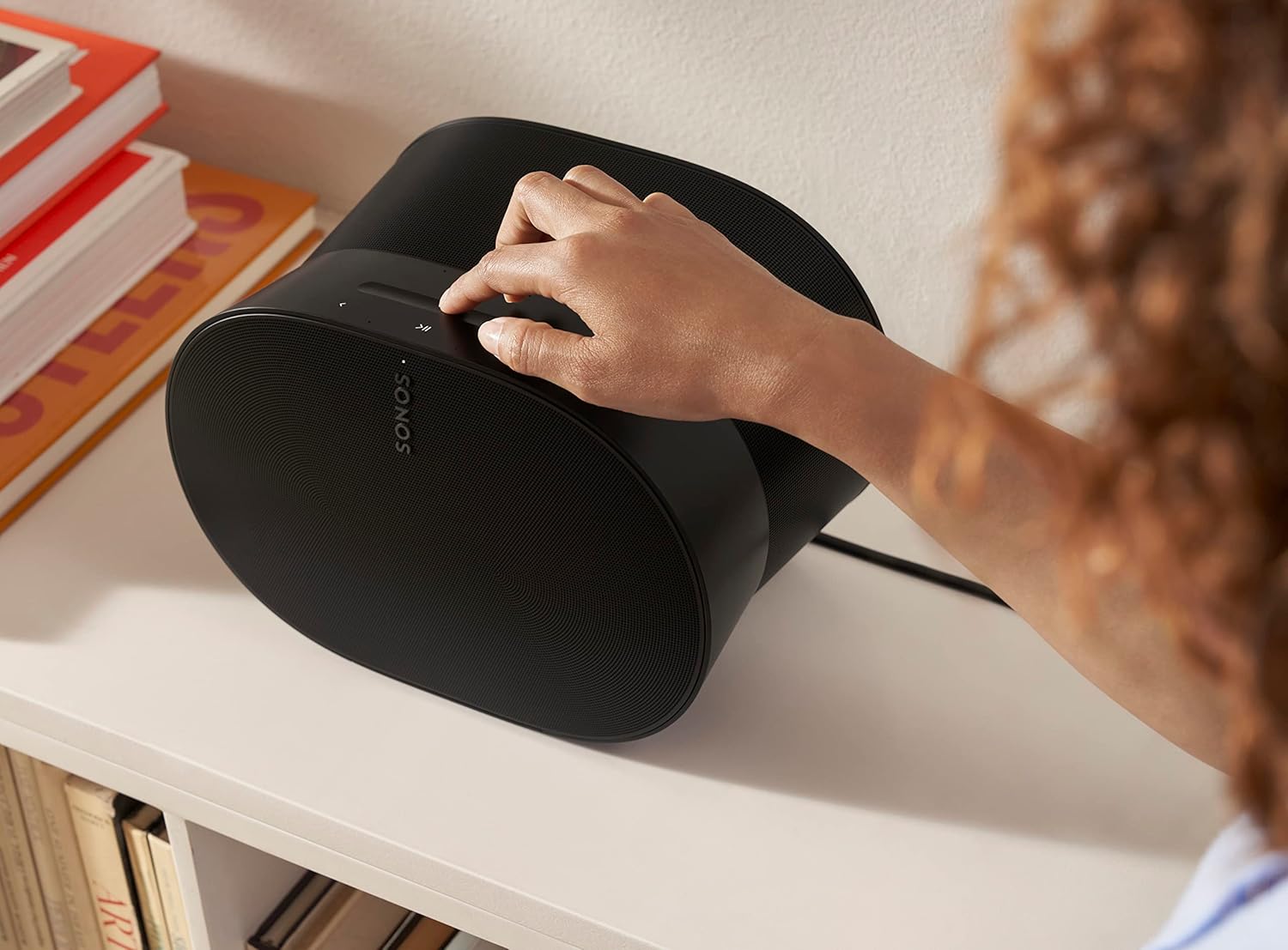 Enceinte Sonos Era 300 en finition noire posée sur un meuble blanc près d'une pile de livres, une main réglant la surface de commande tactile sur le dessus, montrant la silhouette en sablier et le logo Sonos en façade
