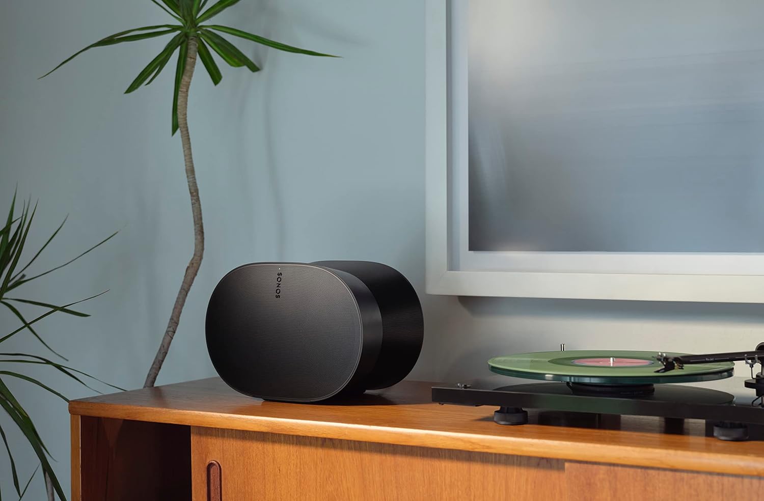 Enceinte Sonos Era 300 en finition noire posée sur un meuble en bois à côté d'une platine vinyle, devant un mur bleu-gris, montrant la silhouette en sablier et le logo Sonos en façade