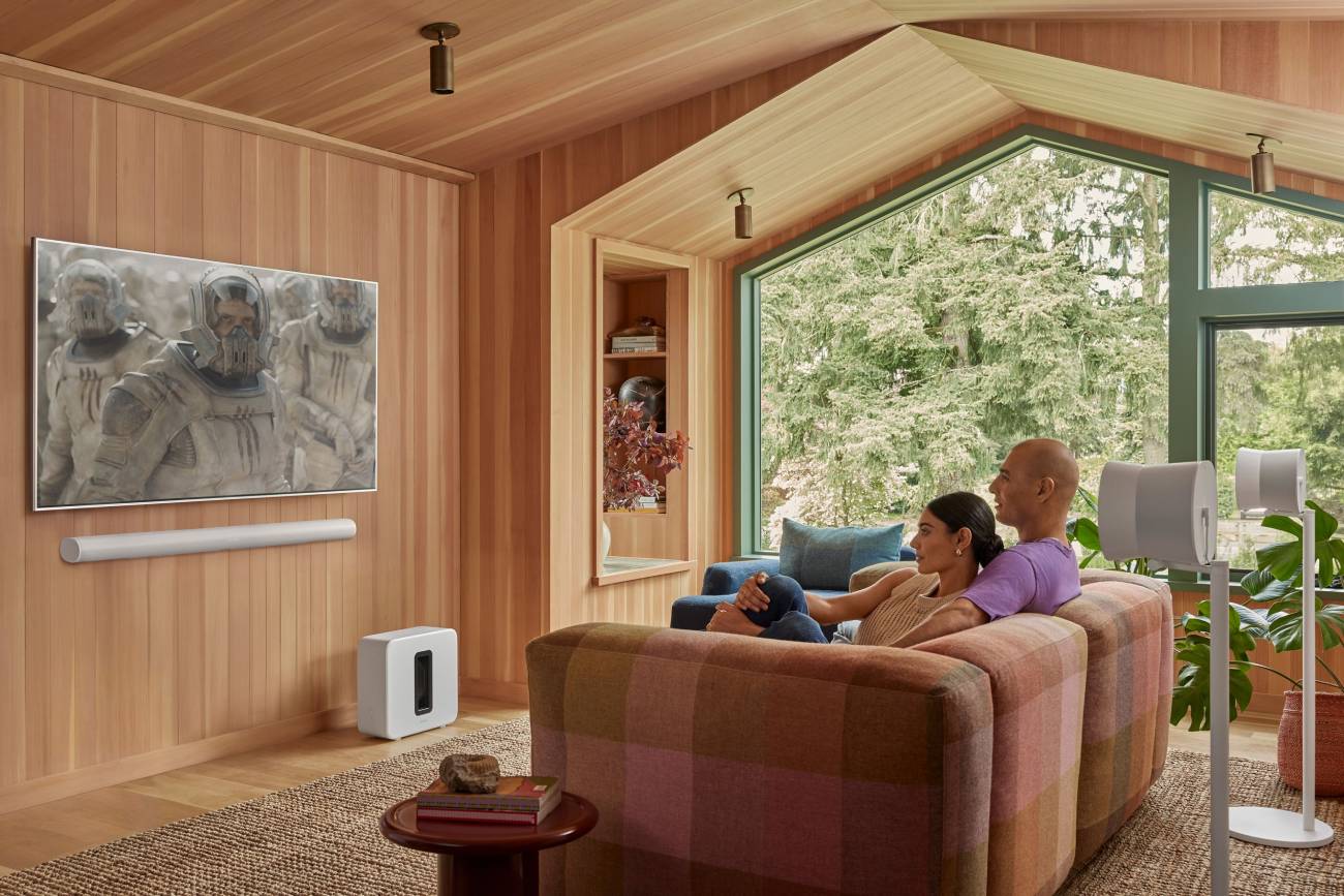 Caisson de basses Sonos Sub 4 blanc posé au sol près d'un meuble, et deux enceintes Era 300 blanches montées sur pieds en position surround derrière un canapé, dans un salon lambrissé de bois clair, illustrant la disposition home cinéma Dolby Atmos du système Sonos