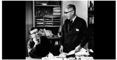Photo d'archive noir et blanc des co-fondateurs de Sony Masaru Ibuka et Akio Morita dans un bureau du siège Tokyo Tsushin Kogyo, l'un assis riant à gauche et l'autre debout consultant des documents techniques, étagères de classeurs et livres en arrière-plan
