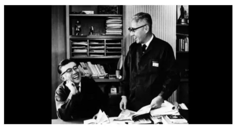 Photo d'archive noir et blanc des co-fondateurs de Sony Masaru Ibuka et Akio Morita dans un bureau du siège Tokyo Tsushin Kogyo, l'un assis riant à gauche et l'autre debout consultant des documents techniques, étagères de classeurs et livres en arrière-plan