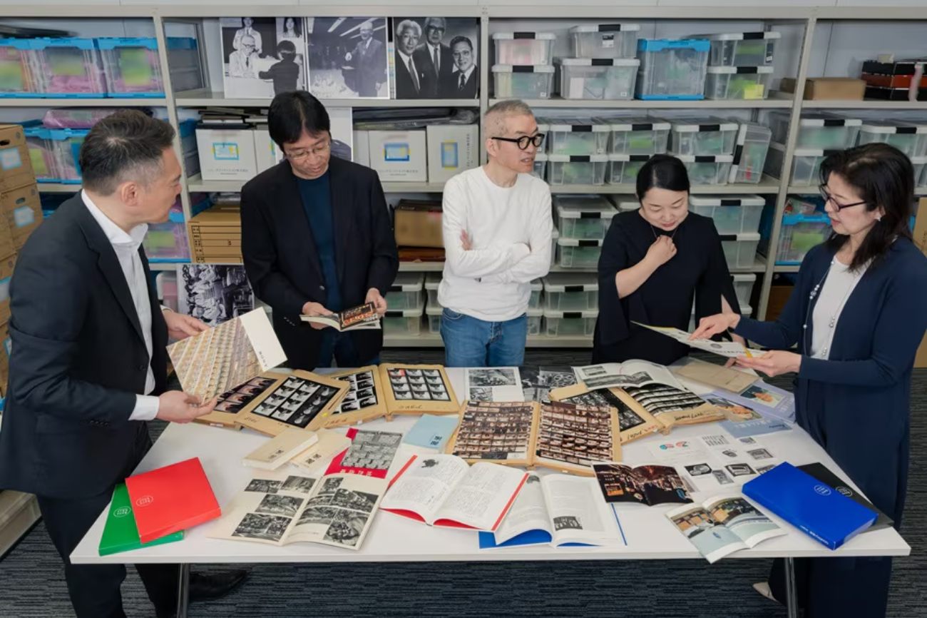 Équipe Archive Team de Sony Group Corporation au travail dans le centre d'archives de Tokyo en 2026, cinq personnes debout autour d'une grande table couverte de photographies argentiques, planches contact, livres techniques d'époque et albums historiques en cours de vérification pour la mise à jour de l'histoire Sony à l'occasion du 80e anniversaire, avec en arrière-plan les rayonnages métalliques garnis de boîtes plastiques translucides d'archives et les portraits encadrés des dirigeants historiques de la marque