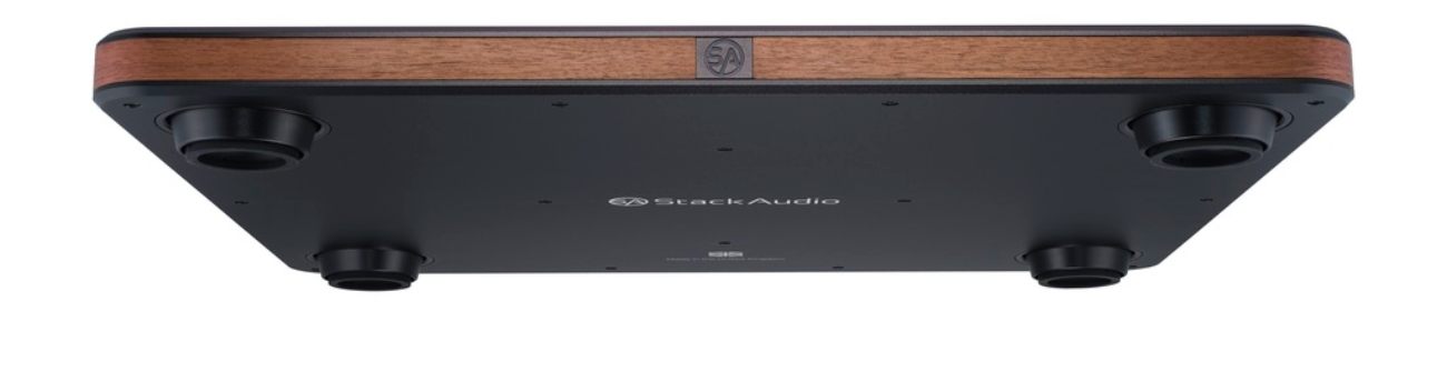 Vue de dessous panoramique de la Stack Audio AUVA Platform sur fond blanc, montrant les quatre pieds CSA Custom Silicone Absorber cylindriques noirs aux quatre coins de la plaque aluminium anodisé inférieure, avec logo Stack Audio gravé au centre et mention Made in United Kingdom