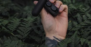 Écouteurs sans-fil SUPRA NERO-TX2 noirs dans leur boîtier de charge Powermag II ouvert, tenus à la main en extérieur sur fond de fougères, illustrant la conception ultra-compacte et la vocation outdoor rugged du modèle
