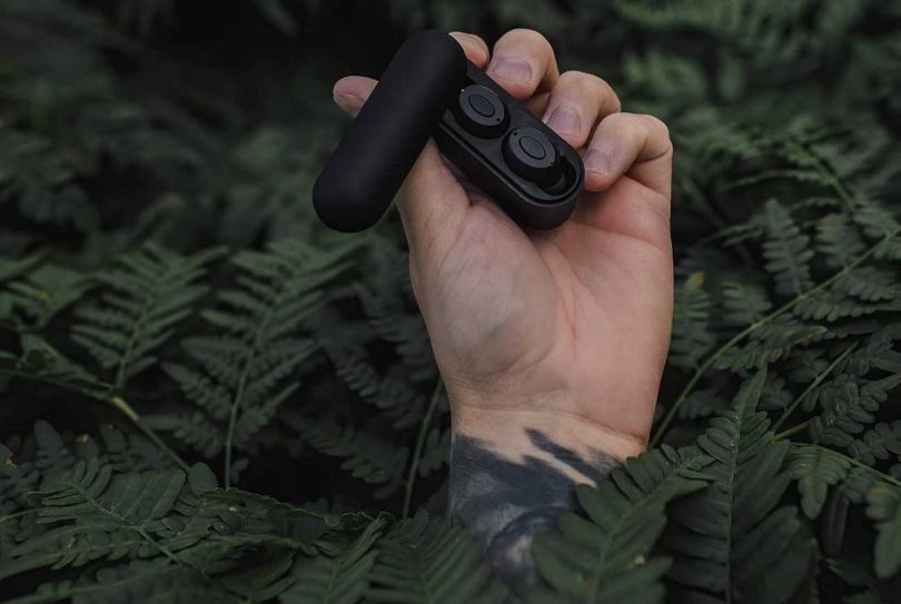 Écouteurs sans-fil SUPRA NERO-TX2 noirs dans leur boîtier de charge Powermag II ouvert, tenus à la main en extérieur sur fond de fougères, illustrant la conception ultra-compacte et la vocation outdoor rugged du modèle
