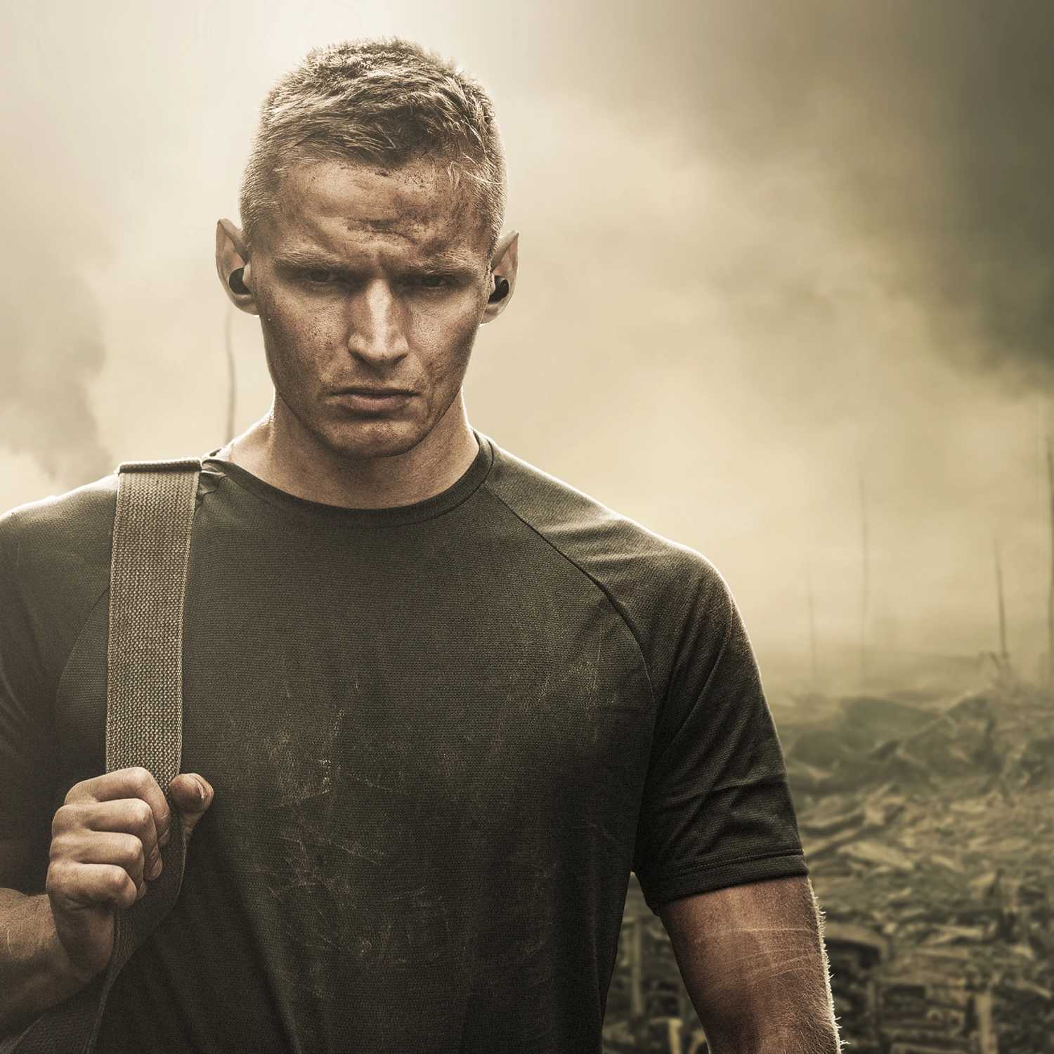 Homme au look militaire, visage marqué et tee-shirt technique, portant un écouteur SUPRA NERO-TX2 à l'oreille dans un environnement extérieur enfumé et accidenté, illustrant le positionnement robuste et la résistance aux conditions difficiles
