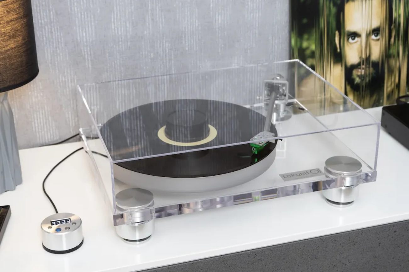 Platine vinyle Takumi TT Level 2.1DC en finition acrylique PMMA transparente avec capot articulé fermé, bras de lecture en titane, plateau acrylique noir, sous-plateau aluminium poli, trois pieds amortissants en aluminium et boîtier de contrôle SpeedPod externe à afficheur numérique posé à gauche, en situation sur un meuble blanc