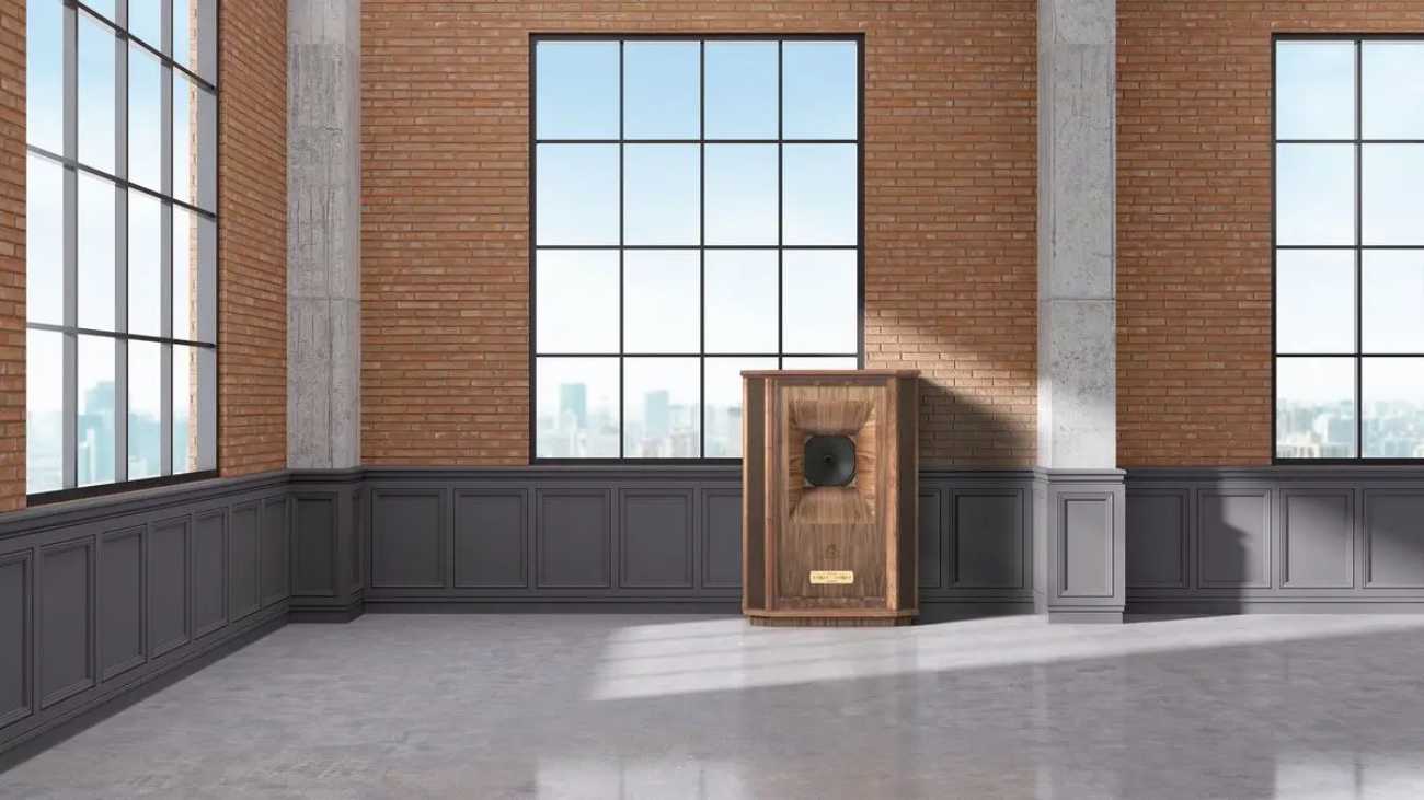 Enceinte Tannoy Canterbury GR édition anniversaire 100 ans en finition noyer huilé avec placage bois en motif papillon autour du haut-parleur Dual Concentric 15 pouces, plaque dorée Tannoy bien visible, mise en scène dans un loft industriel contemporain avec briques rouges et grandes baies vitrées.