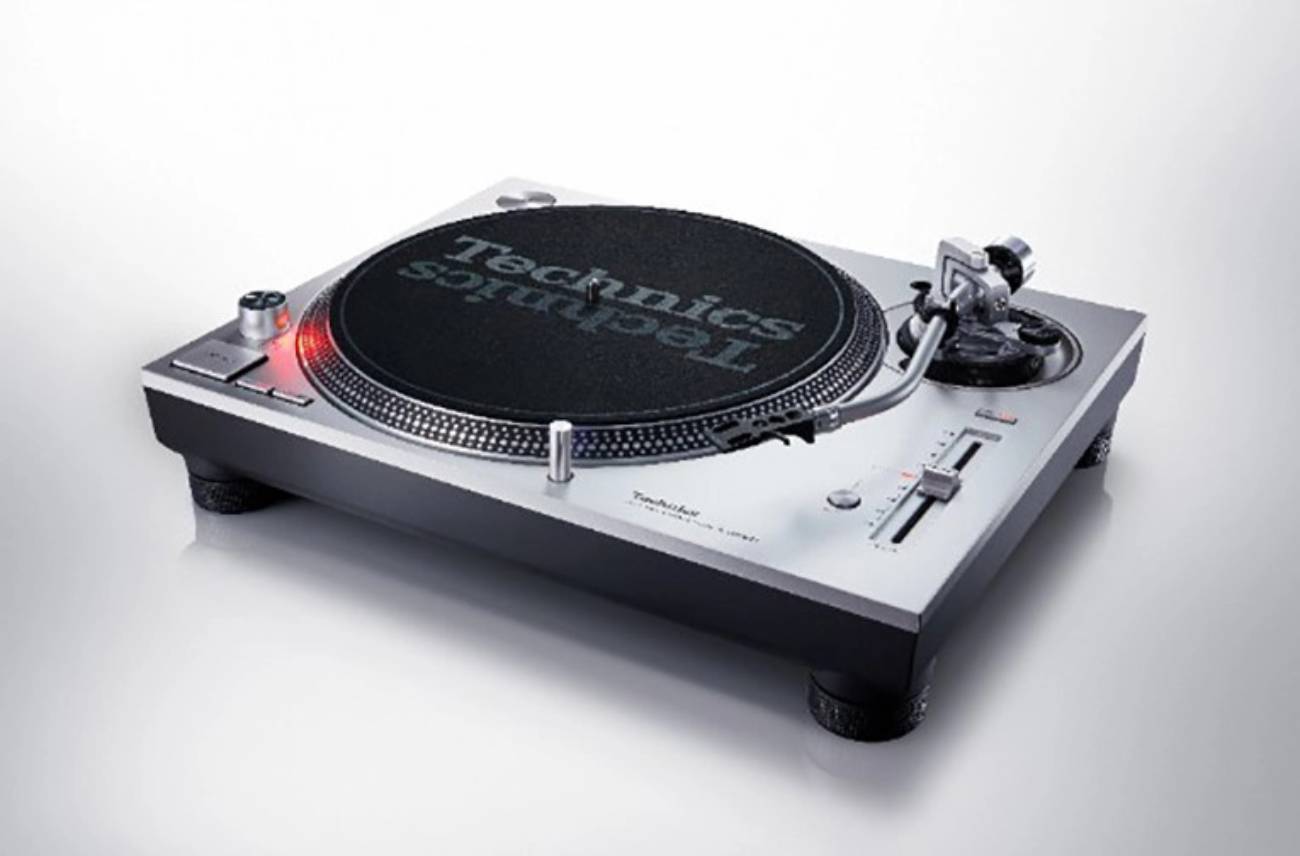 Vue trois-quarts de la platine vinyle Technics SL-1200MK7W en finition argent (référence S), montrant le bras S statique universel avec sa cellule headshell, le levier de pitch avec sa LED stroboscopique rouge allumée, le slipmat Technics noir, et la massivité du châssis bicouche aluminium moulé sous pression posé sur ses quatre pieds amortisseurs.