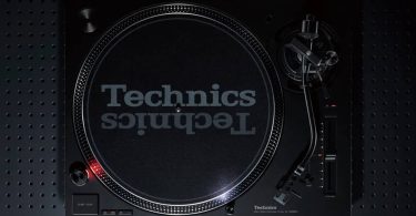 Vue de dessus de la nouvelle platine vinyle Technics SL-1200MK7W en finition noire, montrant le plateau bicouche aluminium avec slipmat Technics, le bras S statique universel à droite avec sa cellule headshell, le levier de pitch avec sa LED stroboscopique, et les commandes 33/45 tours en bas. Posée sur un panneau perforé sombre.
