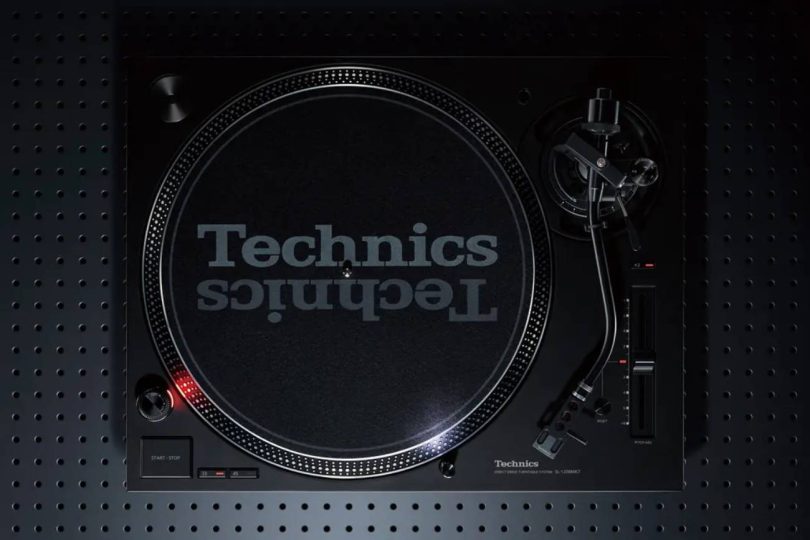 Vue de dessus de la nouvelle platine vinyle Technics SL-1200MK7W en finition noire, montrant le plateau bicouche aluminium avec slipmat Technics, le bras S statique universel à droite avec sa cellule headshell, le levier de pitch avec sa LED stroboscopique, et les commandes 33/45 tours en bas. Posée sur un panneau perforé sombre.