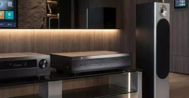 Amplificateur de puissance Tone Winner AD-3240PA noir trois canaux posé sur une étagère en verre dans un salon contemporain, associé à un AVR sur sa gauche et à une enceinte colonne B&W série 600 à sa droite, avec finition murale en bois sombre rétroéclairée