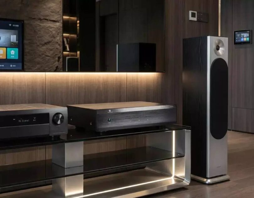 Amplificateur de puissance Tone Winner AD-3240PA noir trois canaux posé sur une étagère en verre dans un salon contemporain, associé à un AVR sur sa gauche et à une enceinte colonne B&W série 600 à sa droite, avec finition murale en bois sombre rétroéclairée