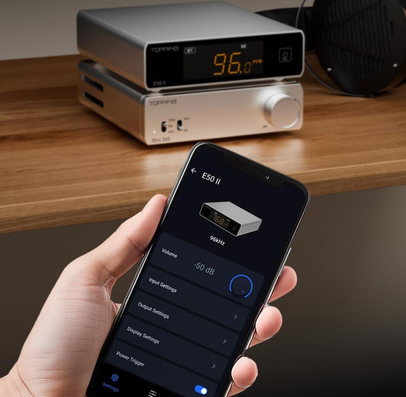Pile composée du DAC TOPPING E50II en finition argent foncé (afficheur orange 96 kHz PCM en mode SE) posé sur l'amplificateur TOPPING Mini300 en finition argent clair, sur étagère en bois massif avec casque audio à droite, et main tenant au premier plan un smartphone affichant l'application Topping Home avec contrôle du volume à -50 dB