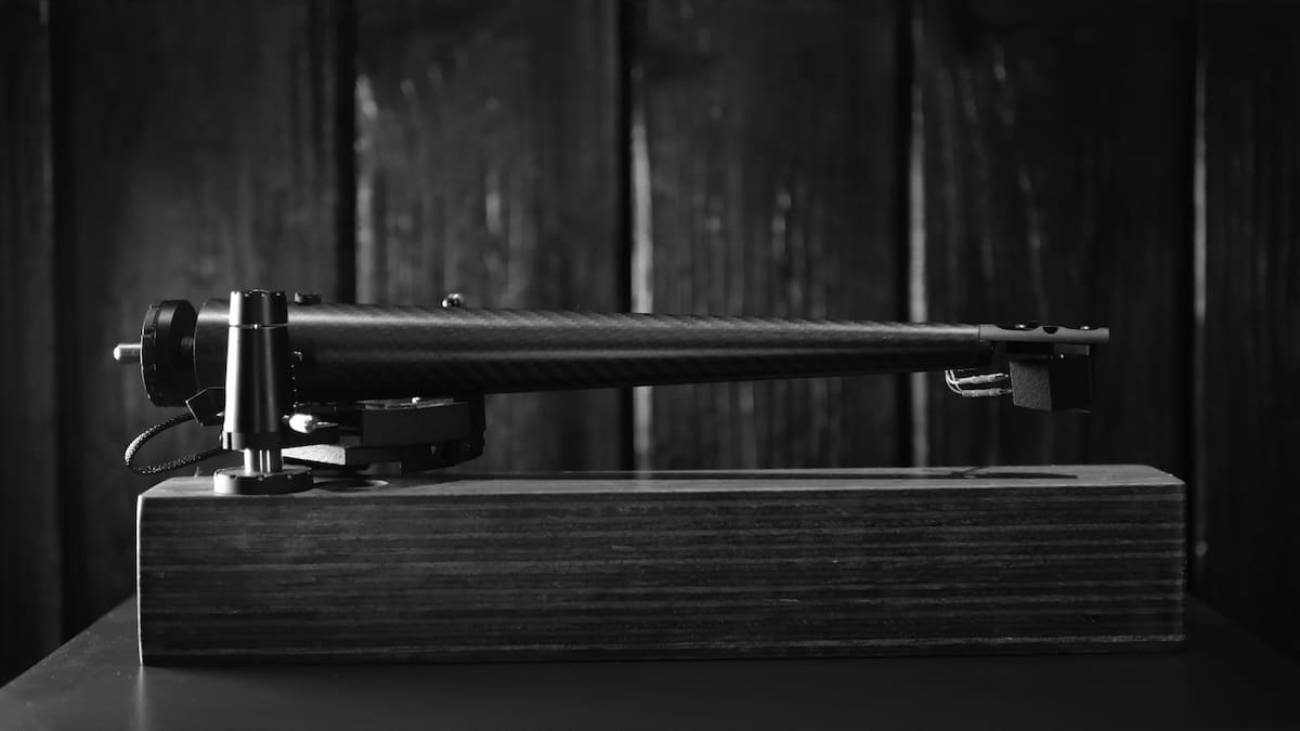 Vue de profil latéral monochrome du bras de lecture Wand Dark-Light 12 pouces présenté sur un socle de bois sombre, tube en fibre de carbone tressée à profil Musical Taper évasé vers le pivot et affiné vers la tête de lecture aluminium, palier ZeroPoint et contrepoids cylindrique noir visibles, ambiance graphique noir et blanc