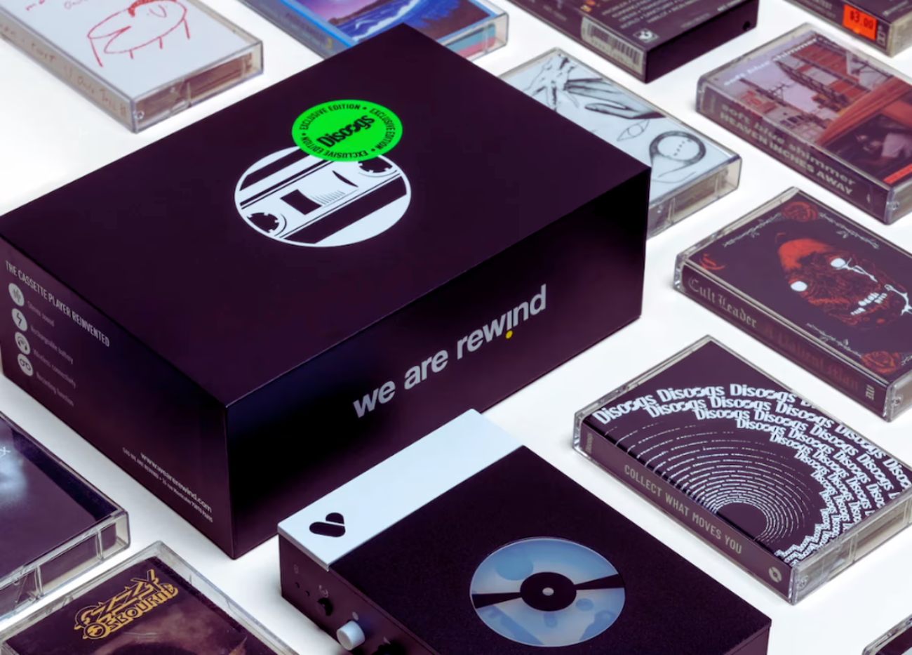 Mise en scène collector du We Are Rewind WE-001 Discogs Edition avec sa boîte de packaging premium noire estampillée logo Discogs vert fluo Exclusive Edition, le lecteur cassette en bas avec logo Discogs visible sur la fenêtre, entouré de cassettes Discogs RTM C60 et de cassettes d'artistes pour illustrer l'univers collector de la plateforme
