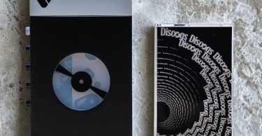 Lecteur cassette portable We Are Rewind WE-001 Discogs Edition en finition Matte Black exclusive avec logo Discogs sur la fenêtre de visualisation cassette, châssis en alliage d'aluminium compact, contrôles de lecture et fenêtre transparente laissant voir la cassette en cours, vue de trois-quarts sur fond neutre