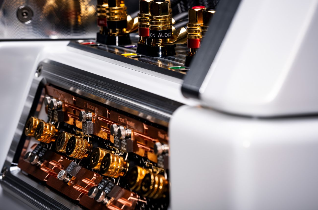 Vue rapprochée du plateau résistances accessible sans outil du crossover de l'enceinte Wilson Audio Autobiography en finition Ethereal White satiné, montrant les résistances cylindriques dorées montées sur un dissipateur en cuivre pur, les étiquettes de routage par voie gravées noir sur cuivre identifiant 2 pouces Midrange, Front Tweeter et Rear Tweeter, le cadre en aluminium machiné argent à arêtes vives qui structure le module, et en arrière-plan flou les bornes de raccordement haut-parleur dorées avec leurs fiches couleur Wilson Audio rouge, blanc, vert, jaune et bleu sur le panneau supérieur de l'enceinte