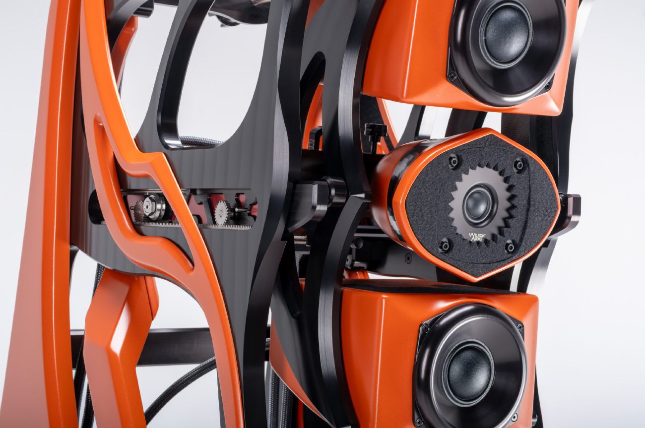 Vue arrière 3/4 du gantry skeletonized de l'enceinte Wilson Audio Autobiography en finition Red Rock Sunset orange, montrant la crémaillère graduée centrale du module alignment sled avec ses deux engrenages crantés argent et son pignon central laqué rouge visible à travers les fenêtres du chariot, les sangles de damping noir mat qui structurent l'arrière du module, le gantry skeletonized en composite aux formes organiques fluides, et l'arrière des trois modules drivers qui composent la face avant : driver médium 7 pouces PentaMag haut, ensemble MTM central, driver PentaMag bas