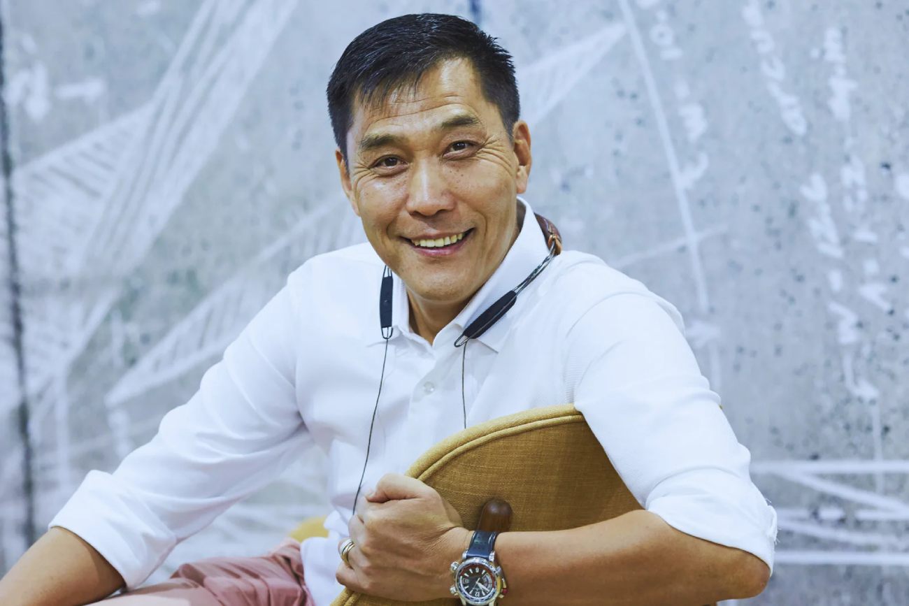 Portrait de Xiaodong Yang CEO de Cinemaster et nouveau propriétaire de Jamo via Jamo Holding Limited souriant en polo blanc avec montre de plongée et écouteurs sport accrochés au cou, sur fond de mur graphique aux motifs techniques en filigrane