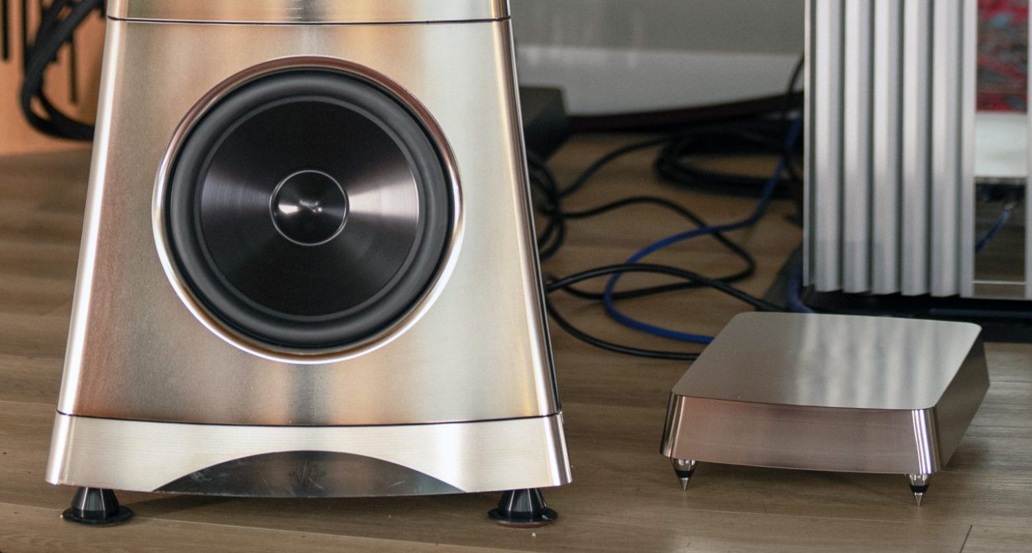 Vue rapprochée de la partie inférieure d'une enceinte YG Acoustics Titan en finition aluminium poli, montrant le woofer sub-bass 32 cm à cône aluminium avec son saladier massif et sa suspension périphérique caoutchouc, la pyramide à pieds spikes en bas, et à droite l'amplificateur dédié 1 kW de la version Active Sub posé sur ses propres pointes isolantes