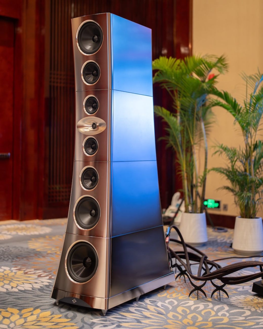 Vue de trois-quarts face d'une enceinte YG Acoustics Titan en finition aluminium teinté cuivre-bronze, montrant l'array vertical de 7 drivers à cônes aluminium et le woofer sub-bass 32 cm en partie basse, présentée en salon de démonstration audiophile avec moquette à motifs floraux et palmiers d'ambiance