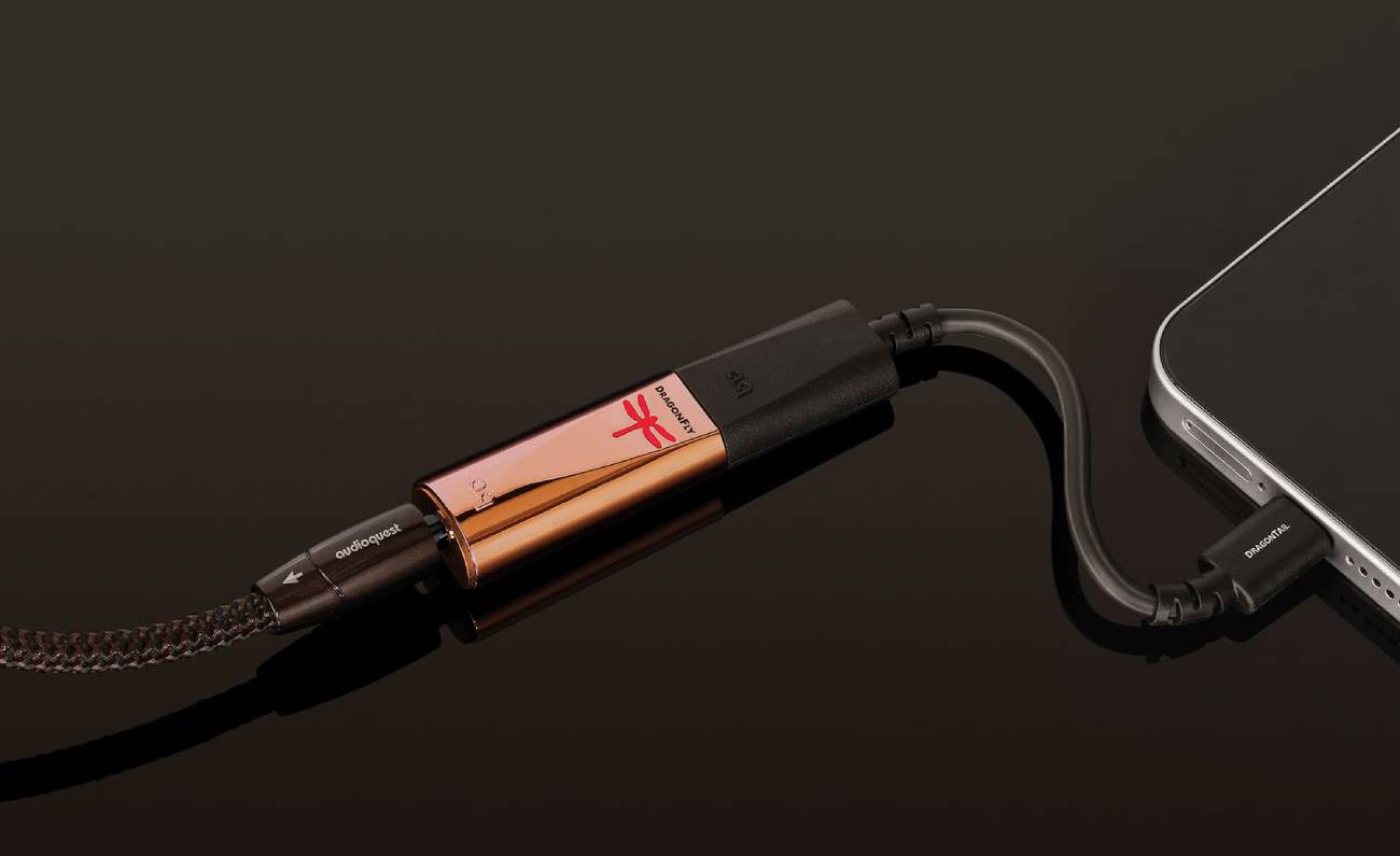DAC USB nomade AudioQuest DragonFly Copper au boîtier cuivre avec logo libellule rouge allumé, relié par l'adaptateur DragonTail USB-C à un smartphone, un câble AudioQuest gainé branché sur sa sortie casque 3,5 mm, sur fond sombre